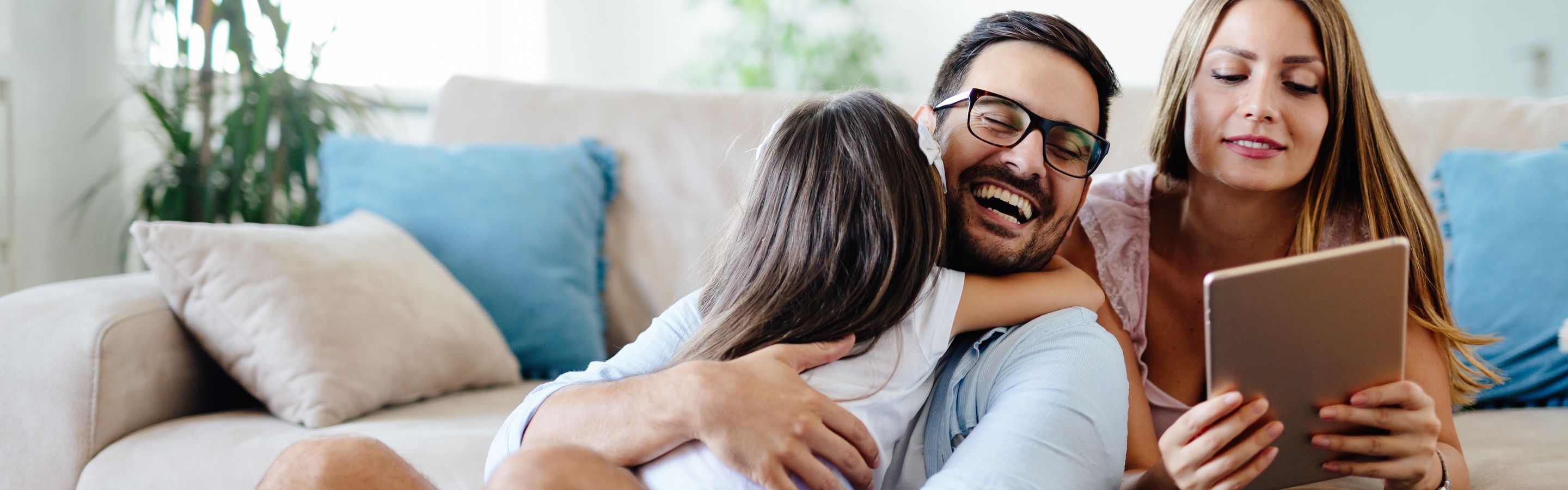 Junges Paar sitzt lächelnd auf Sofa, der Vater sein Kind im Arm, die Mutter lesend